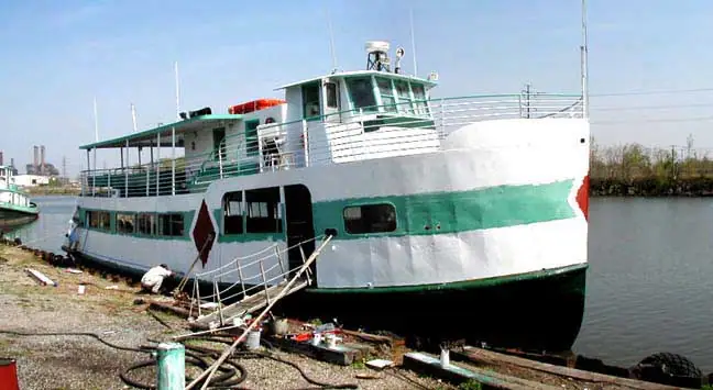 White-and-green ferry moored beside a waterfront work area, with a gangway, hoses, and equipment in the foreground.