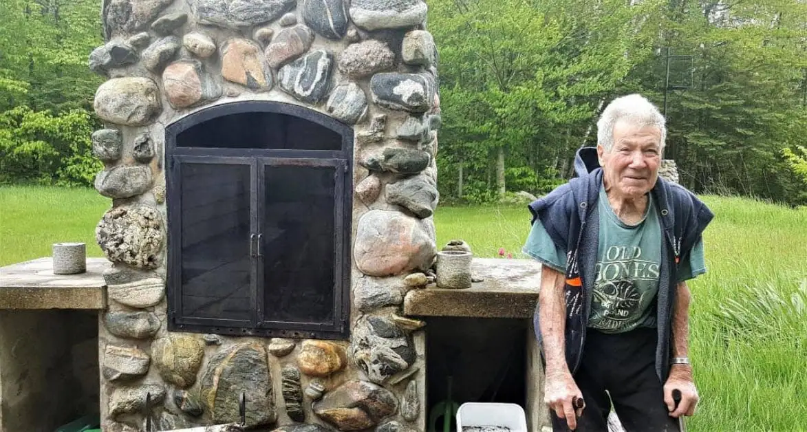 An older man stands beside a large fieldstone outdoor fireplace in a grassy clearing.