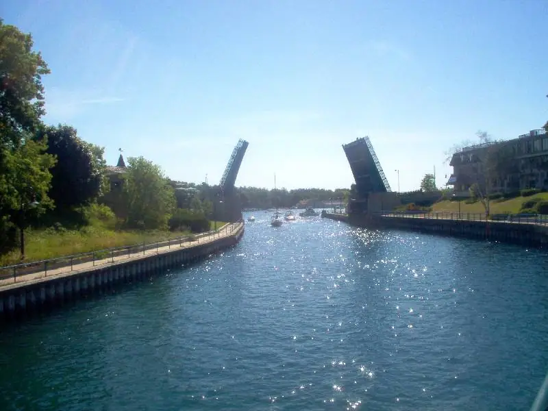 A water channel leads toward two raised drawbridge spans, with boats, walkways, trees, and waterfront buildings.