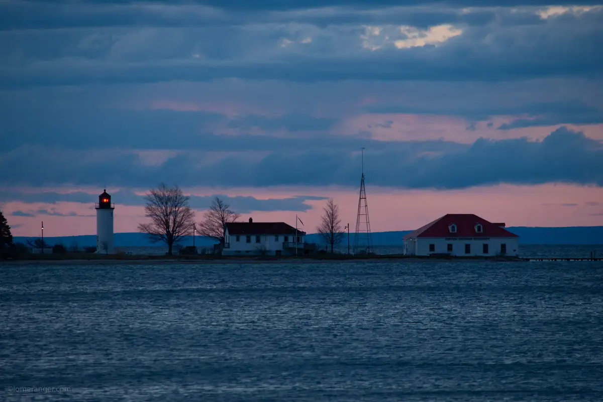 A lighthouse and waterfront buildings stand against a pink and blue twilight sky.