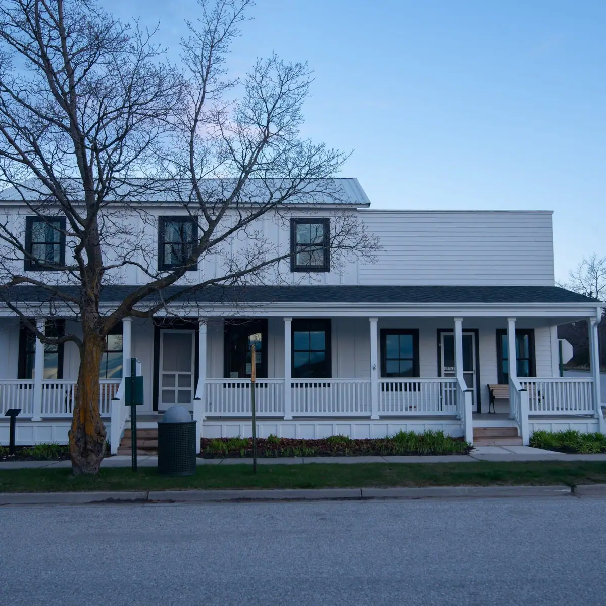 A white two-story building with a covered porch stands behind a leafless tree beside a paved street.