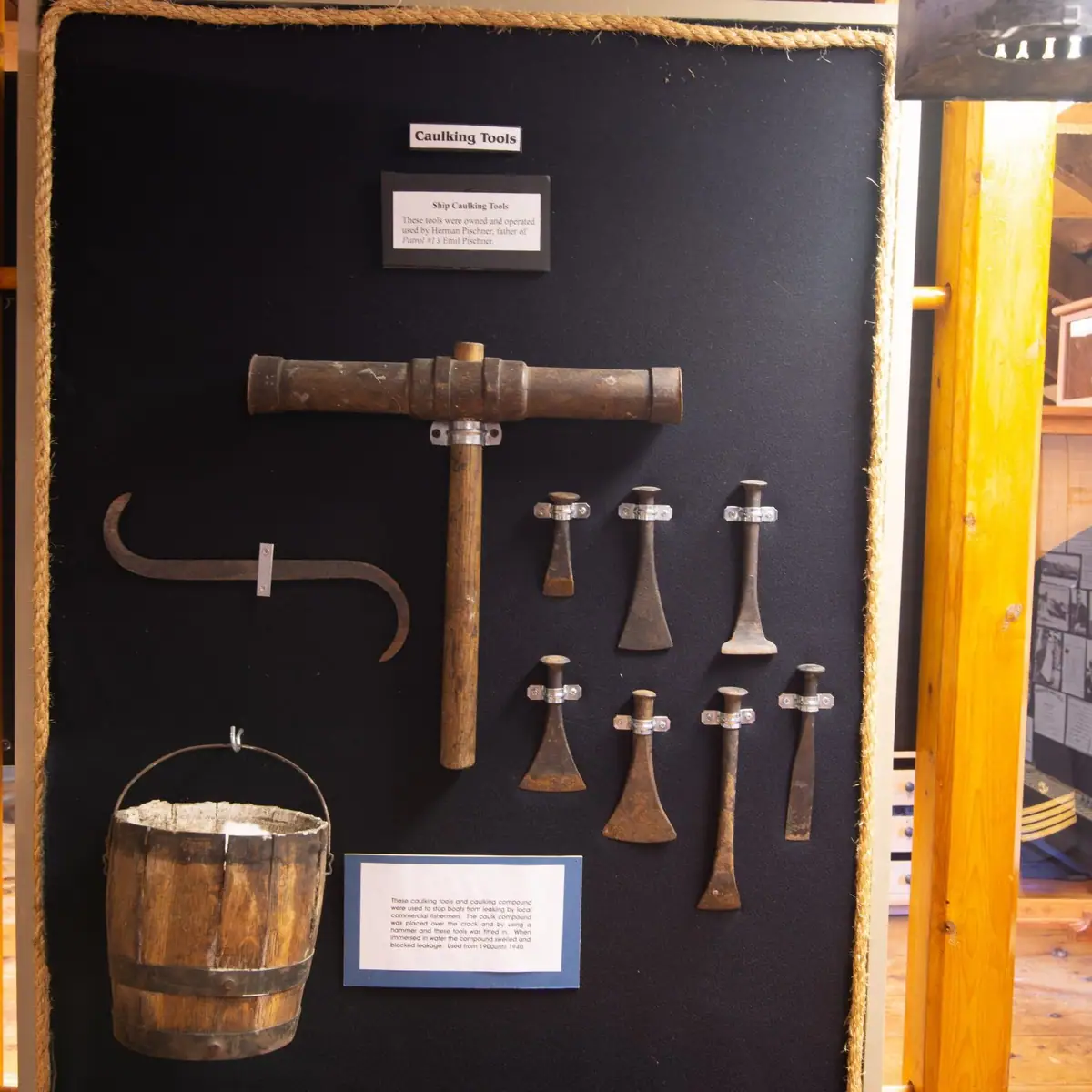 Museum display of ship caulking tools, including a wooden mallet, metal tools, and a wooden bucket.