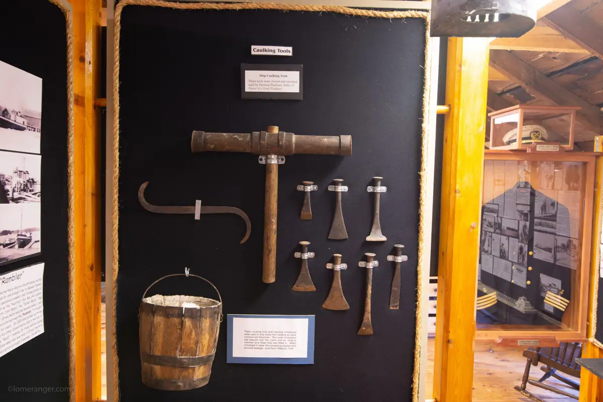 Museum display of caulking tools, including a wooden mallet, metal tools, and a wooden bucket.