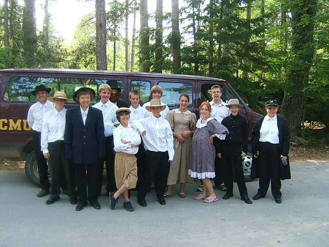 A group in period-style clothing poses beside a maroon bus in a wooded setting.