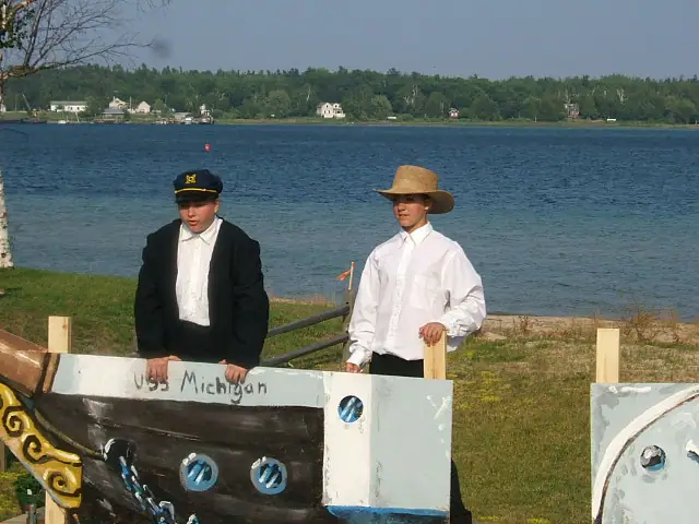 Two costumed young men stand behind a painted ship model beside the water.
