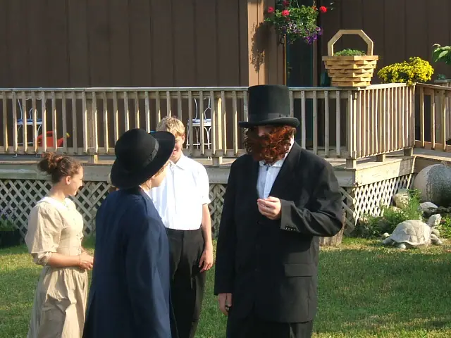 Four people in period-style clothing stand in a yard beside a porch.