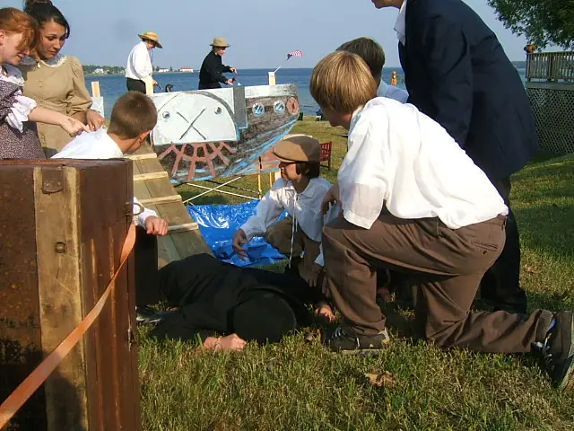 Adults and children gather around a painted wooden boat-shaped prop beside the water.