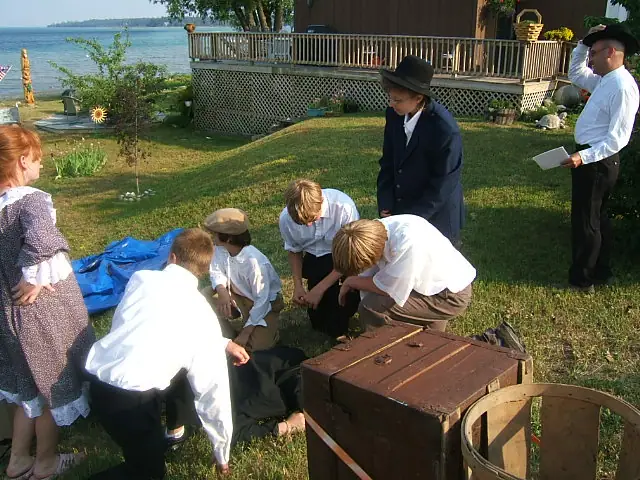 Children and adults in period clothing gather around a wooden trunk in a grassy yard beside the water.