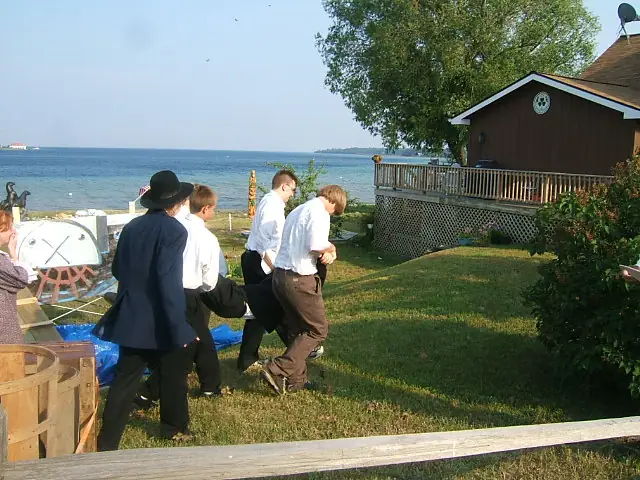 Five men walk across a grassy yard beside a broad body of water, with a house and deck behind them.