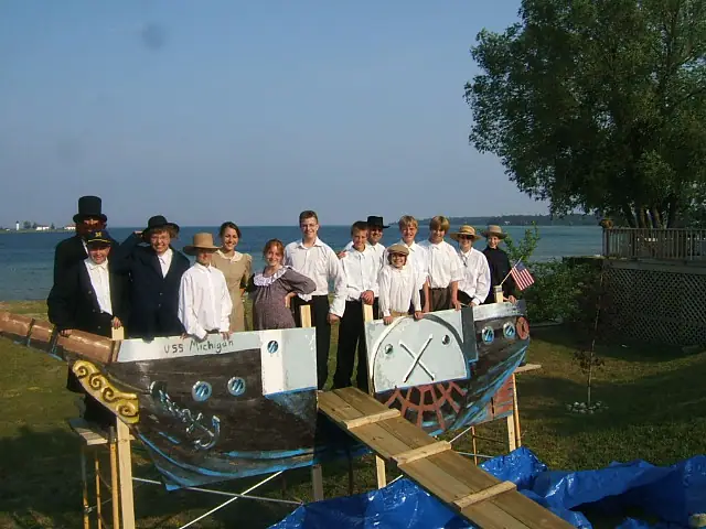A group in period-style clothing poses behind a painted wooden boat model beside the water.