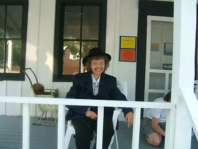 A smiling person in a dark suit and hat sits on a white porch beside a partially visible seated person.