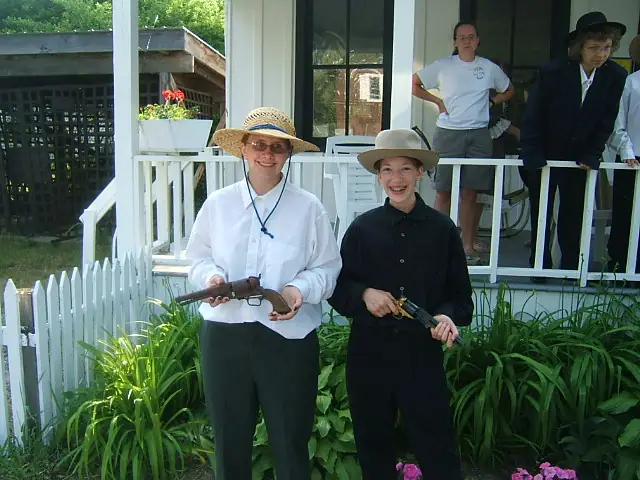 Two people in period-style clothing hold firearms on a house porch while others stand behind them.