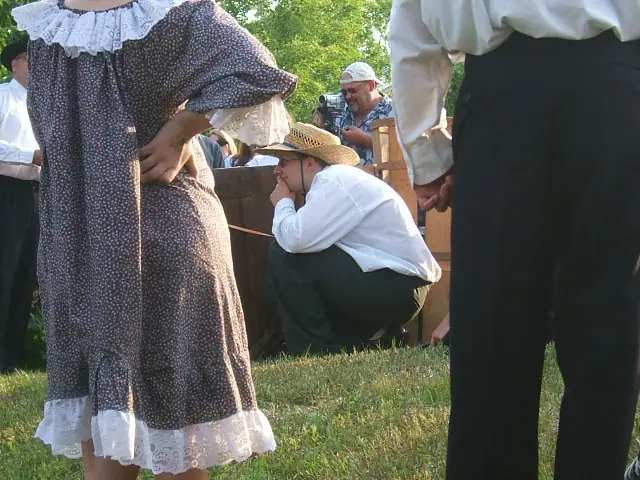 People in period-style clothing gather outdoors beside a wooden enclosure.