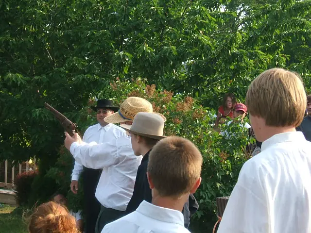 A man holds a long gun while adults and children in brimmed hats watch in a leafy outdoor setting.