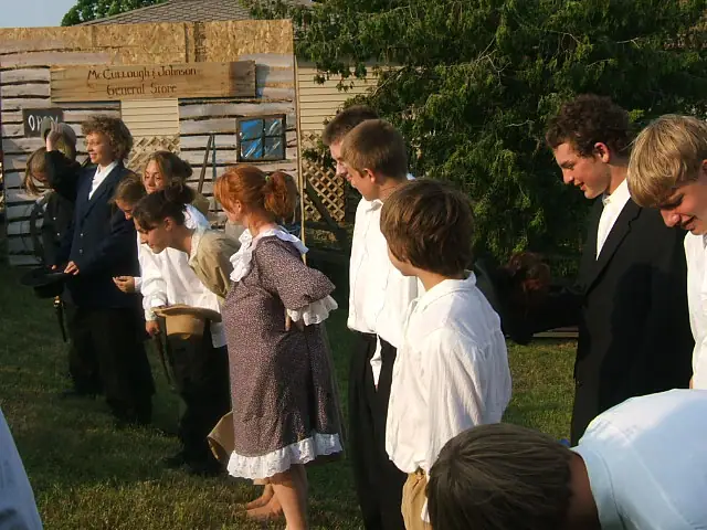 Young people in period clothing stand beside a log building with general-store signs.