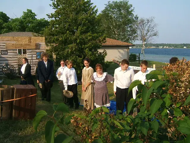 Several adults and children in historical clothing stand beside a wooden storefront and waterfront.