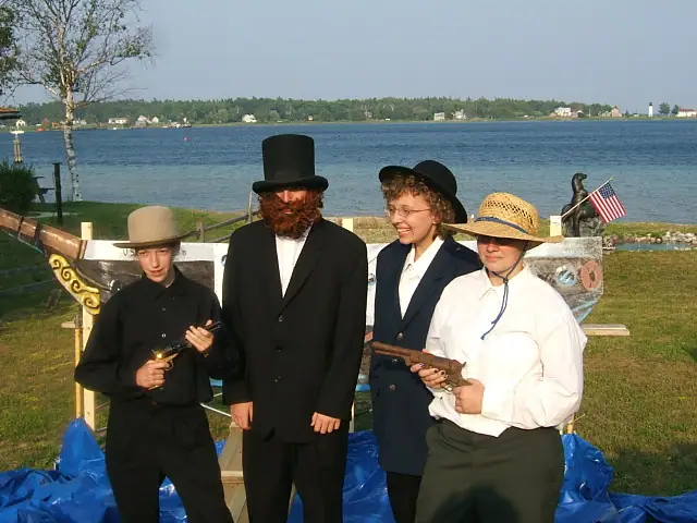 Four people in historical-style clothing stand beside a decorated wooden boat near the water.