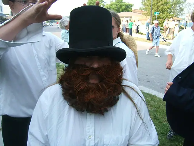 A person in a tall black hat and large reddish beard stands among people on a sunny street.
