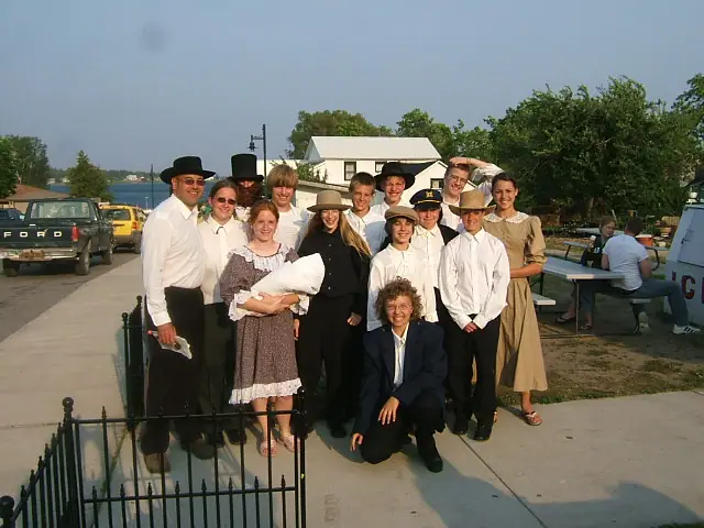 A posed group in period-style clothing stands outdoors beside a paved walkway and waterfront.
