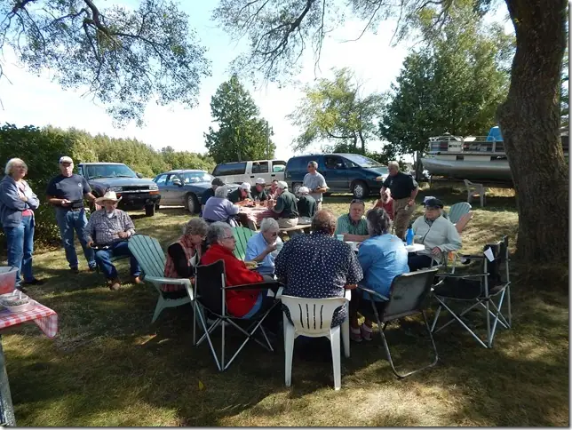 Adults gather around outdoor tables beneath trees, with parked vehicles and a small boat behind them.