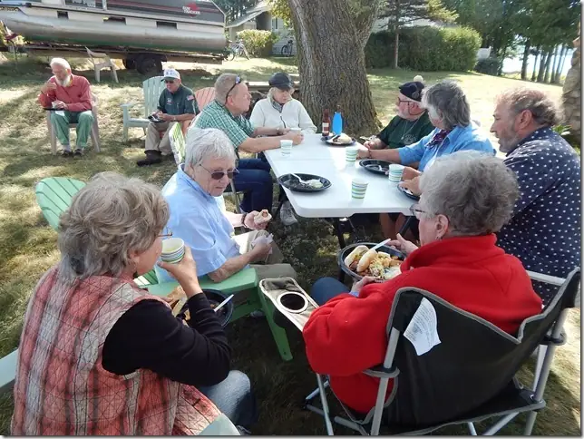 Adults share a meal at a folding table beneath a tree, with a boat on a trailer behind them.