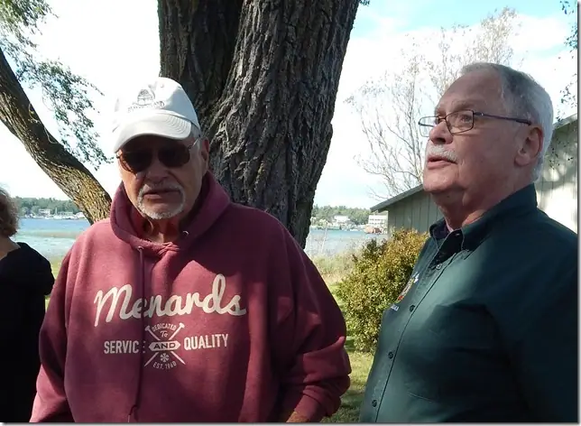 Two men stand beside a large tree with a waterfront and buildings behind them.