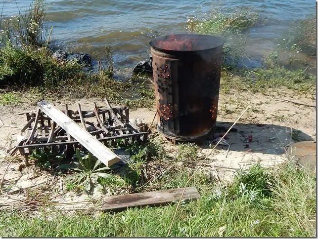A perforated metal barrel stands beside scattered wood and debris on a sandy, grassy shoreline.
