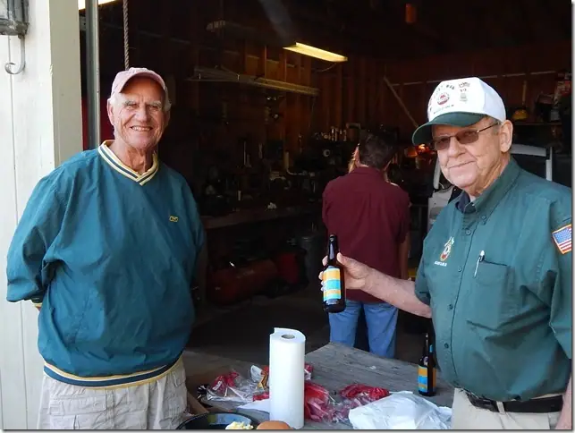 Three people gather at the entrance to a workshop, with one holding a bottled drink beside a food-covered table.