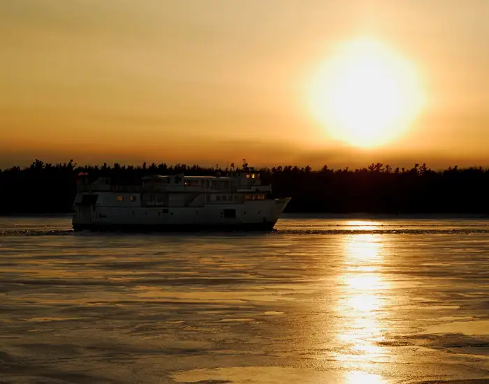 A white ferry crosses reflective ice-covered water beneath a low golden sun and dark tree-lined shore.