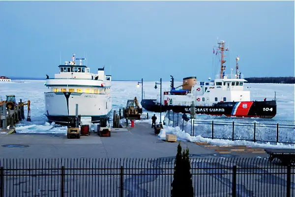 Two vessels sit beside a pier amid broken ice, including a white ferry and a Coast Guard cutter.
