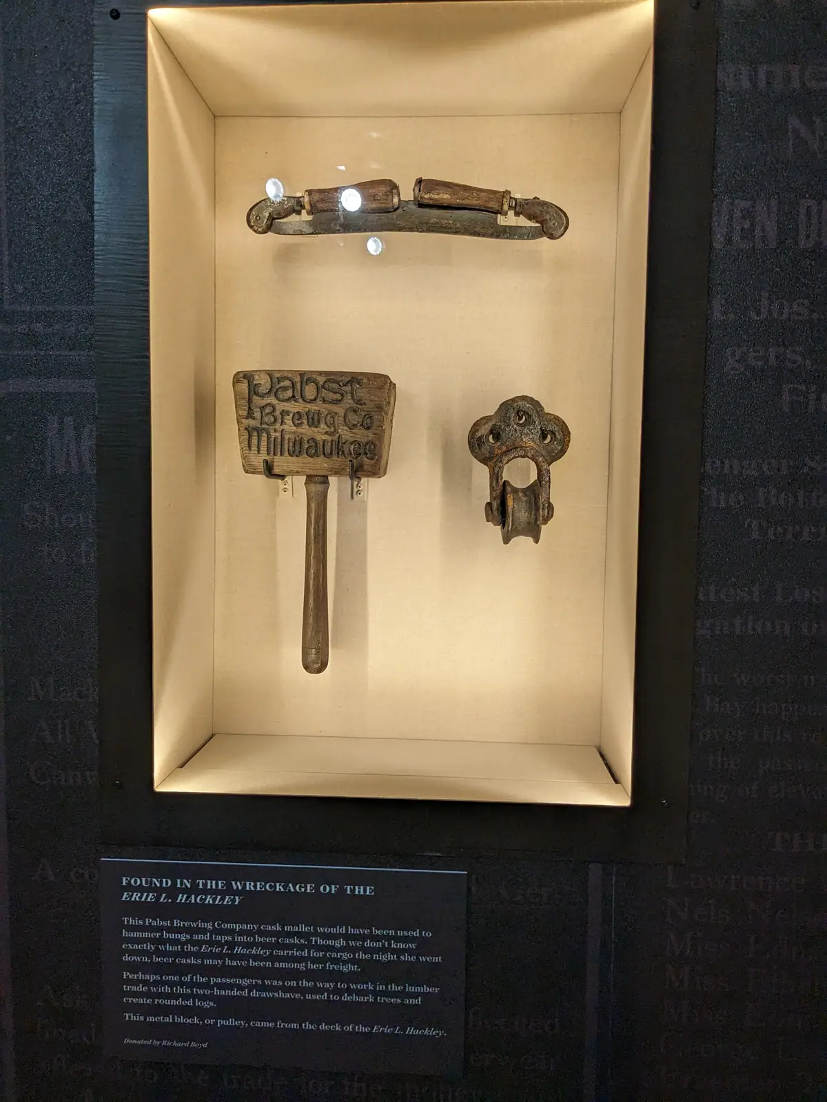 A museum display case holding three objects recovered from the wreck of the Erie L. Hackley: a two-handled drawshave, a wooden cask mallet stamped 'Pabst Brewg Co. Milwaukee', and a corroded metal deck pulley block, with a placard captioned 'FOUND IN THE WRECKAGE OF THE ERIE L. HACKLEY.'
