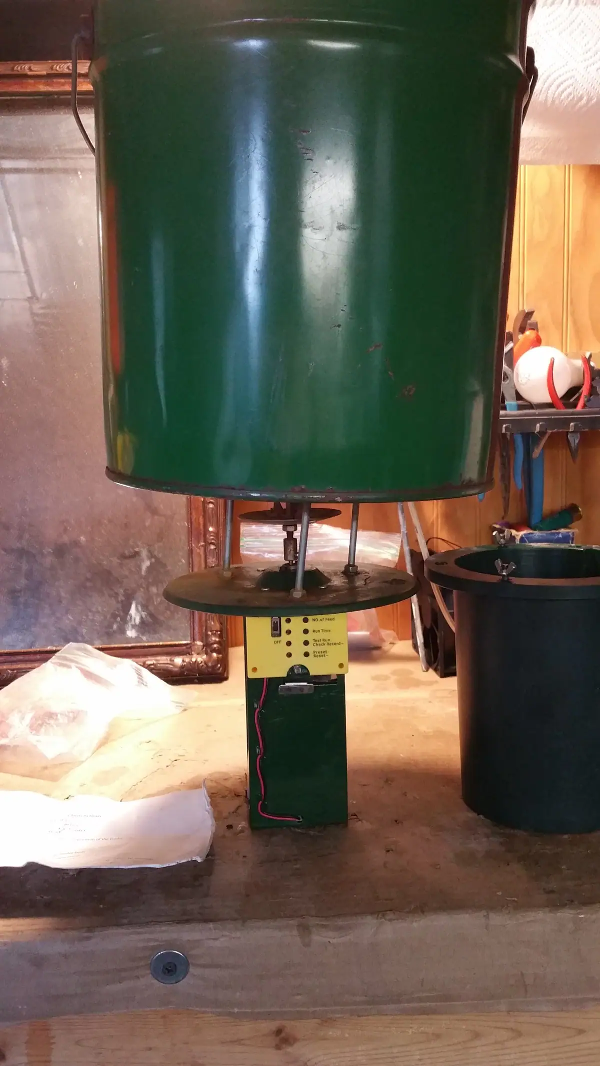 A large green cylindrical feeder with a yellow control panel stands on a mechanical support in a workshop.