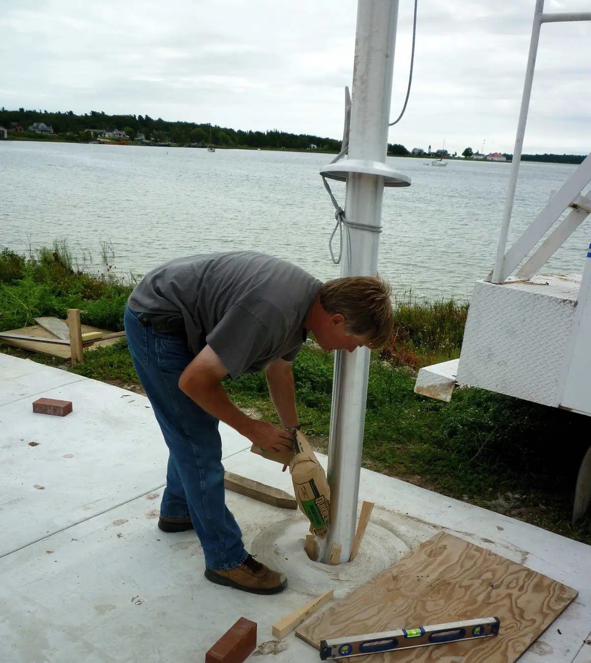 A worker pours material around a metal pole on a concrete platform beside the water.