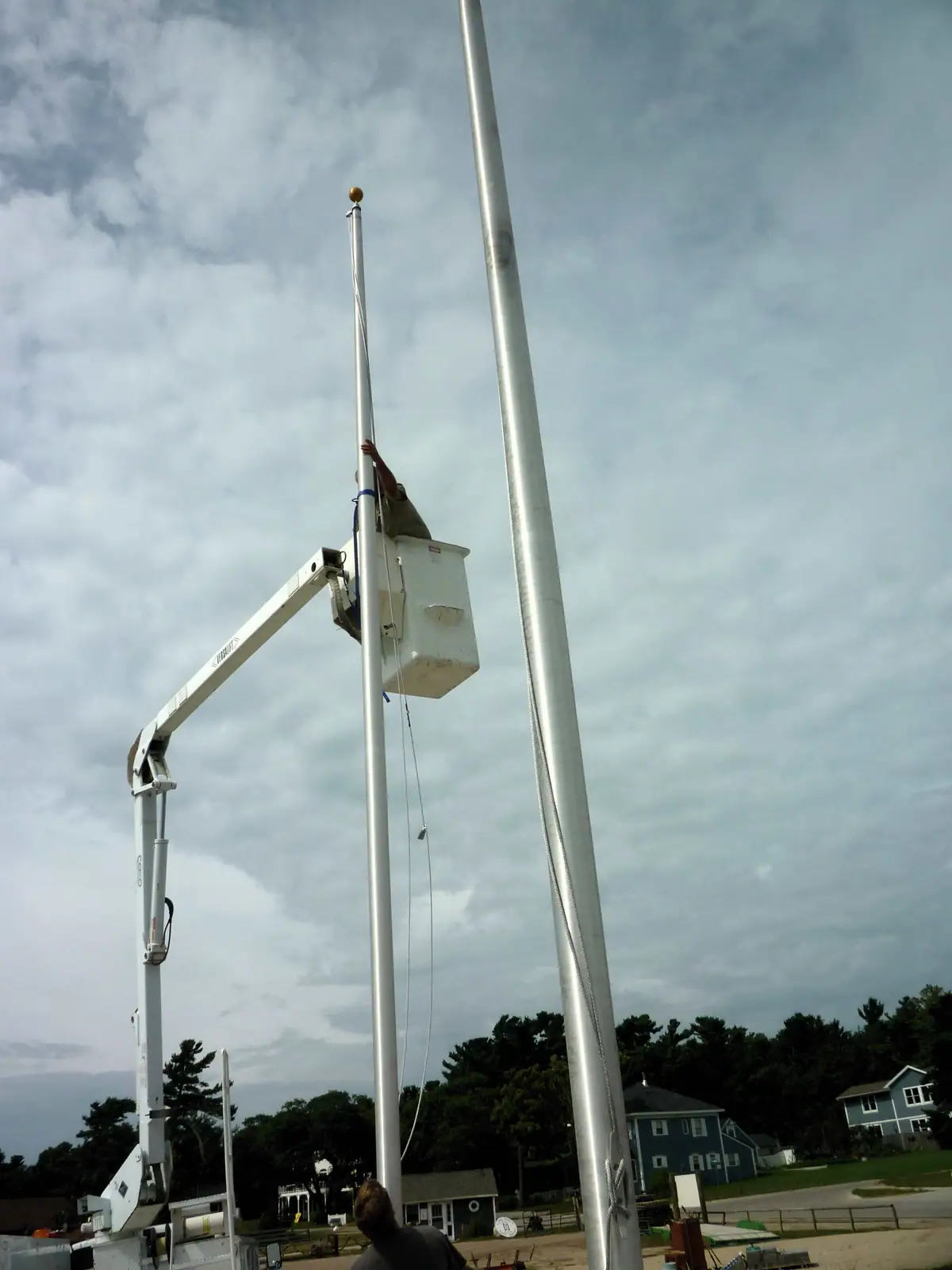 A worker in a raised bucket reaches toward a flagpole beside a second tall pole and a residential street.