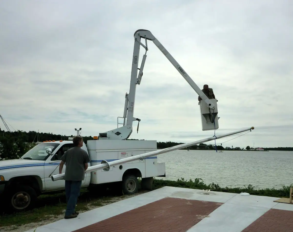 A boom lift truck and two workers handle a long pole beside the waterfront.