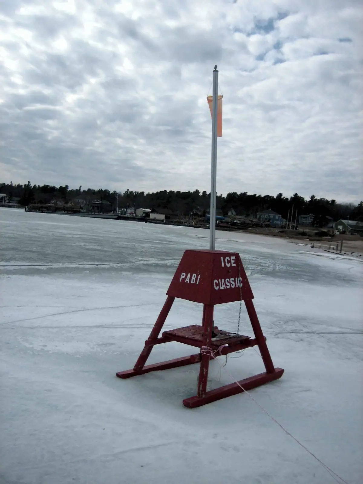 A red metal marker with a pole and flag stands on frozen water near a wooded shoreline.