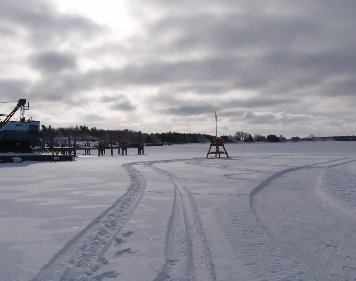 Snow-covered frozen waterfront with tire tracks, a dock, heavy equipment, and a flagged red frame.