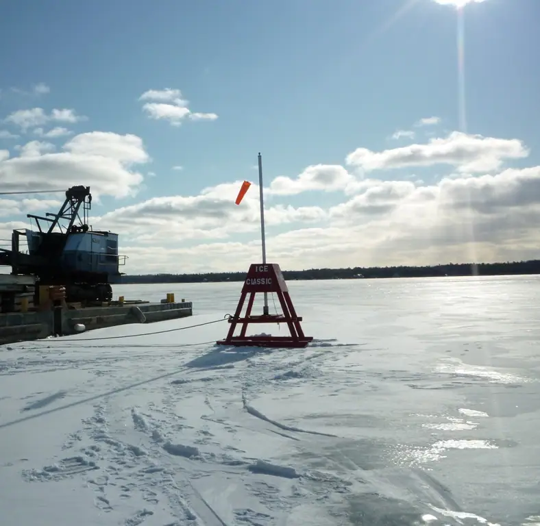 A red ICE CLASSIC marker stands on a snowy frozen expanse beside a crane and dock.