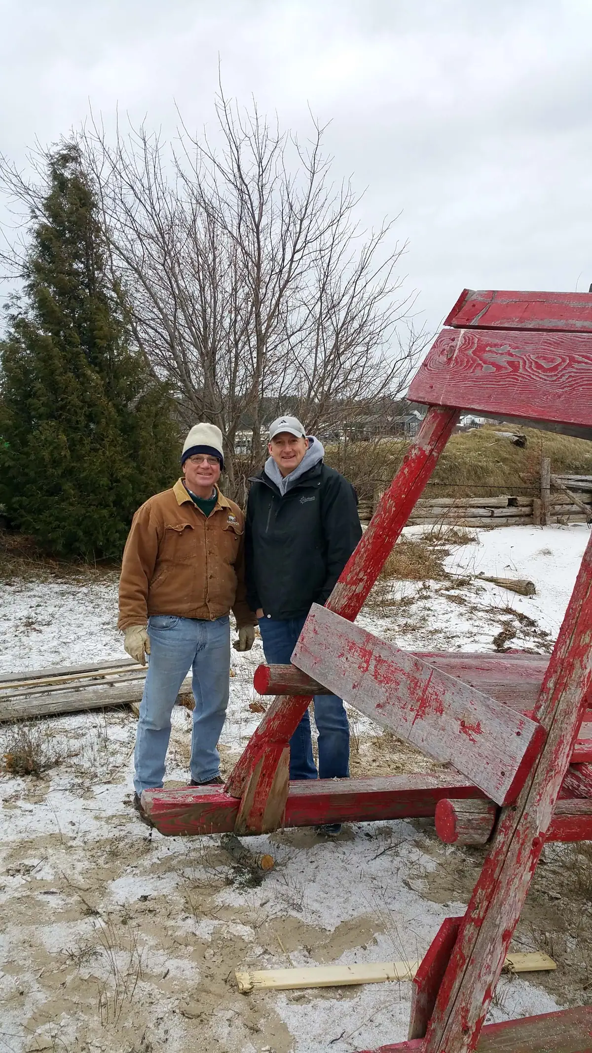 Two men stand beside a weathered red wooden frame in a lightly snow-covered outdoor area.
