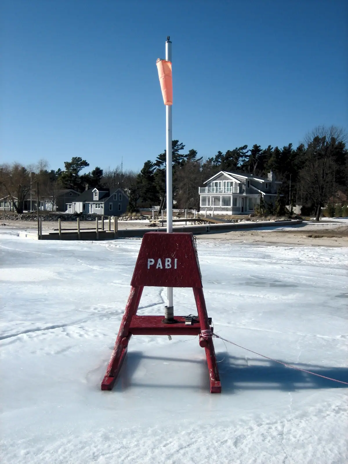 Red A-frame marker labeled PABI stands on ice before waterfront houses and a dock.