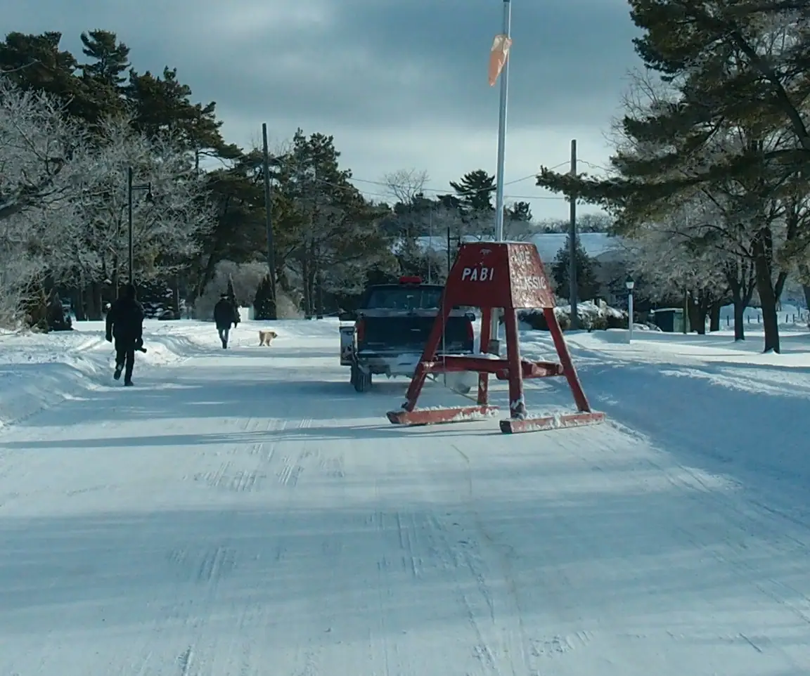 Snow-covered road with a red A-frame sign, parked pickup truck, flagpole, pedestrians, and frosted trees.