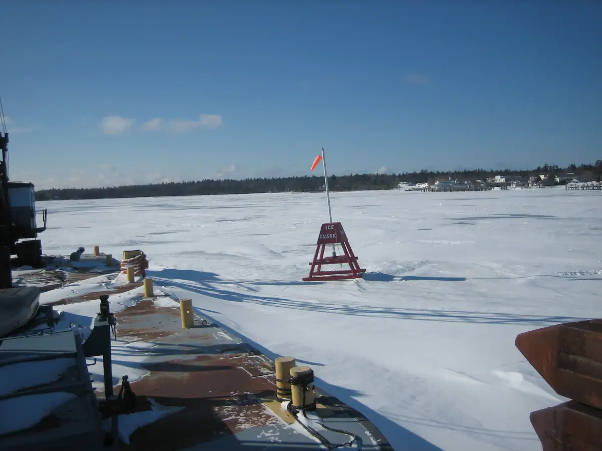 A red ICE CLASSIC marker stands on snow-covered ice beside a vessel deck, with shoreline homes and trees beyond.