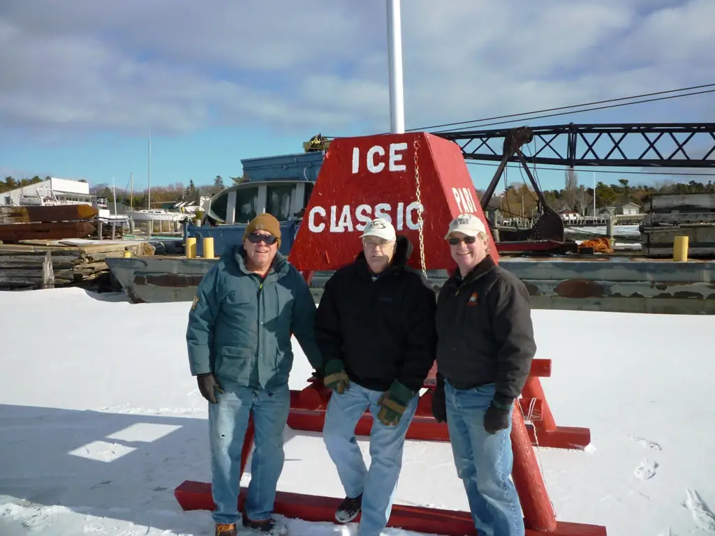 Three men stand in winter clothing beside a red structure marked “ICE CLASSIC” near boats and docks.