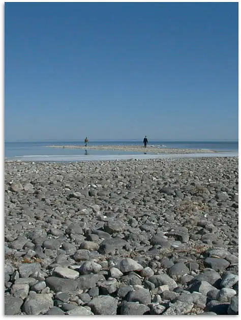 Two people walk along a narrow, pale strip of shore extending into calm water beyond a field of stones.