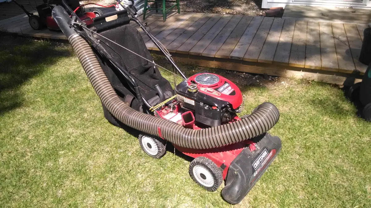 A red walk-behind lawn mower with a large flexible vacuum hose rests on a grassy lawn.