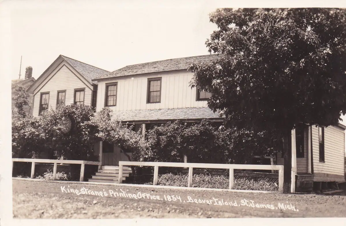 Two-story wooden building with a covered porch, trees, shrubs, and a white fence.
