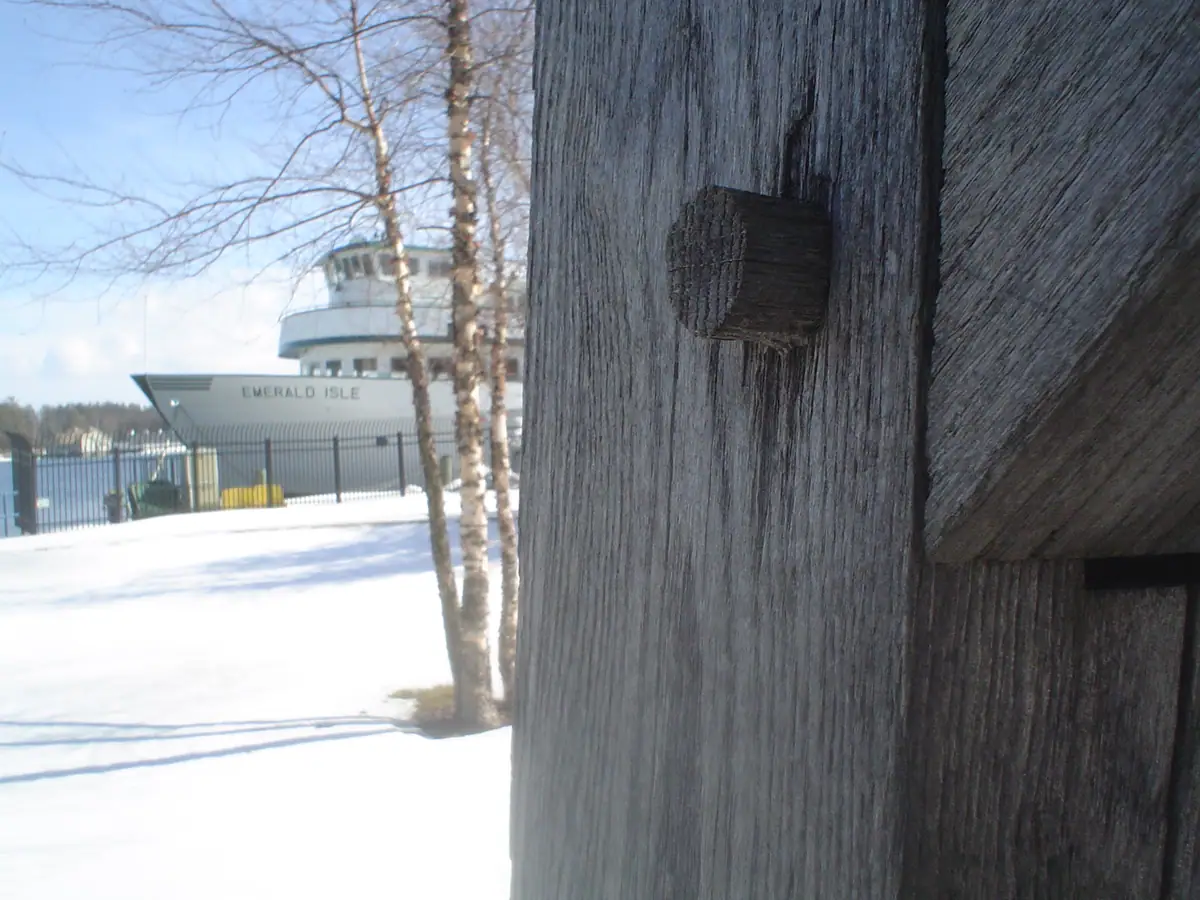 A white ferry marked “EMERALD ISLE” stands behind birch trees and a weathered wooden structure in snow.