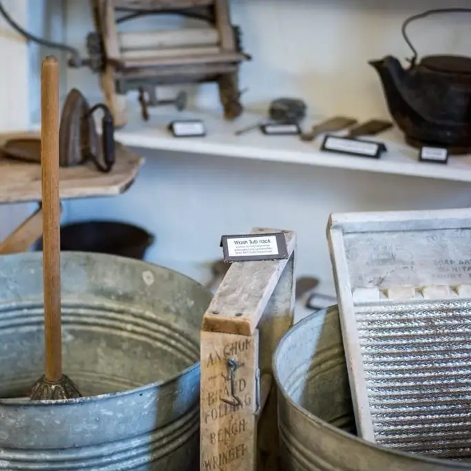 A museum display of galvanized wash tubs, a wooden wringer, a washboard, and other household tools.