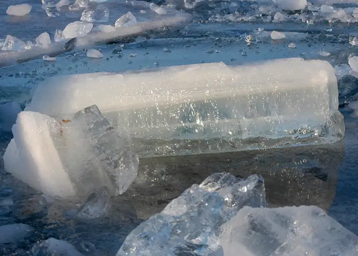 A large layered block of translucent ice lies among smaller fragments on a wet, reflective surface.
