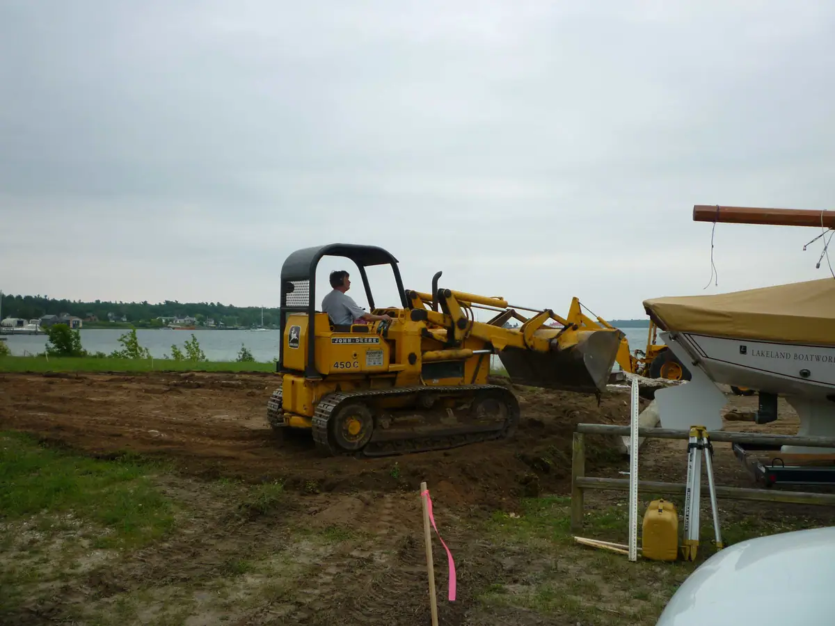 A tracked loader works beside a covered boat on a trailer near the waterfront.
